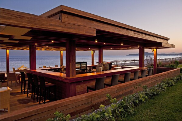 Zona de bar exterior de madera con asientos, con vistas a un cuerpo de agua.