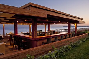 Zona de bar exterior de madera con asientos, con vistas a un cuerpo de agua.