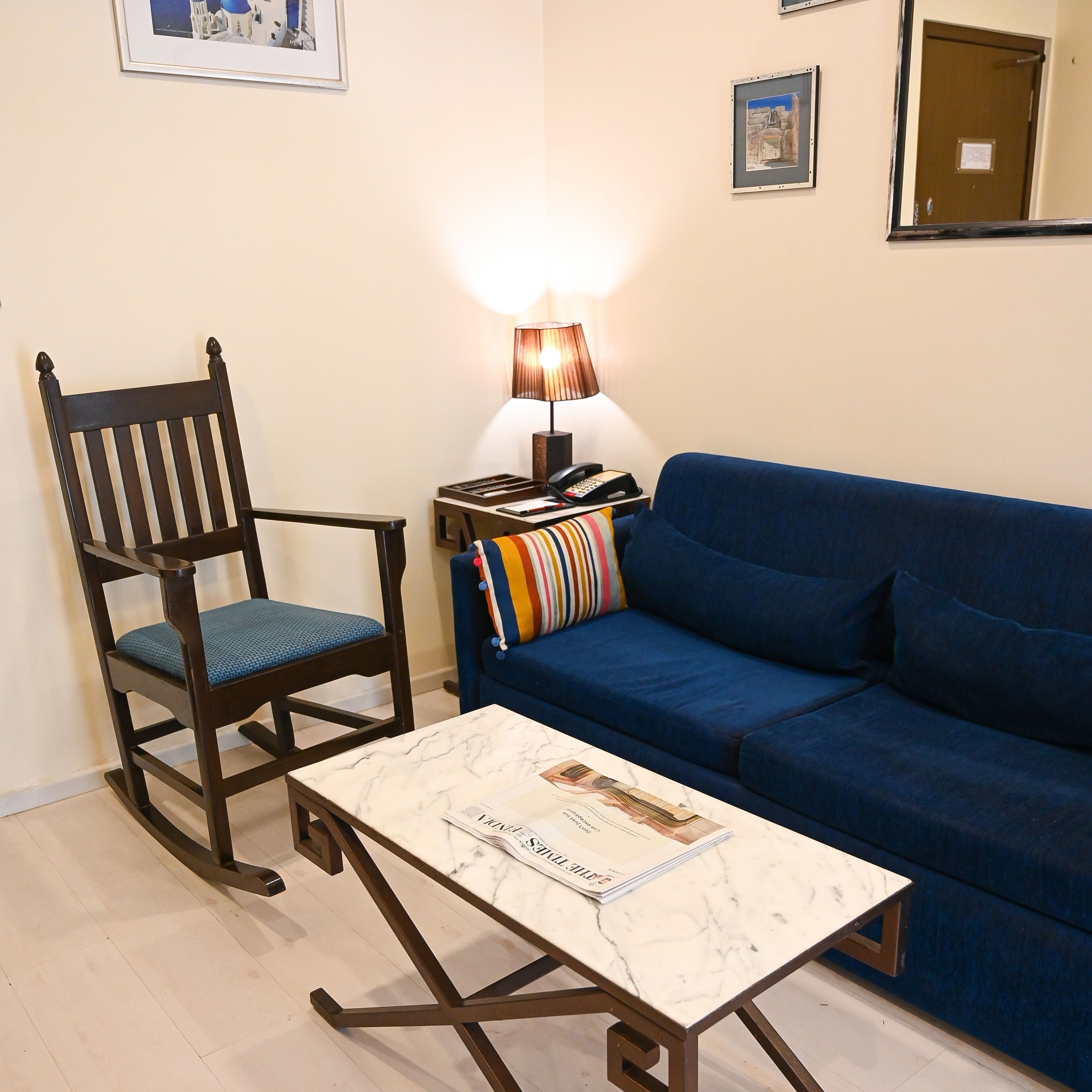 Uma sala de estar com uma cadeira de madeira, um sofá azul, uma mesa pequena com uma revista e uma luminária em um criado-mudo.
