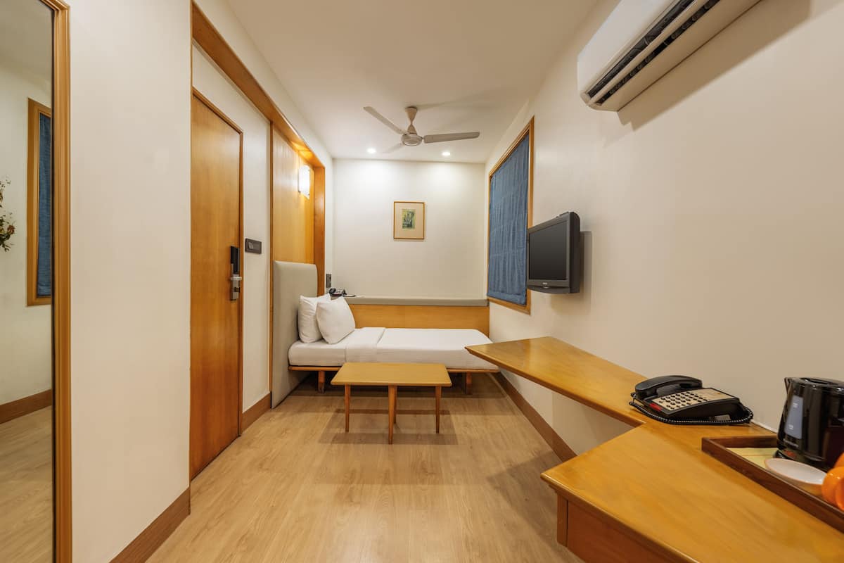 A hotel room with a wooden floor, a sofa, a small table, a desk with a telephone and a coffee maker, a ceiling fan, and an air conditioning unit.