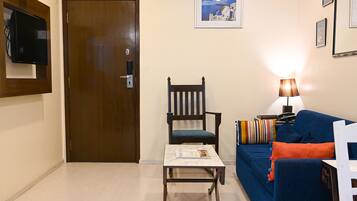 A living room with a wooden door, a blue sofa, a wooden chair, a small table, and a wall-mounted TV.