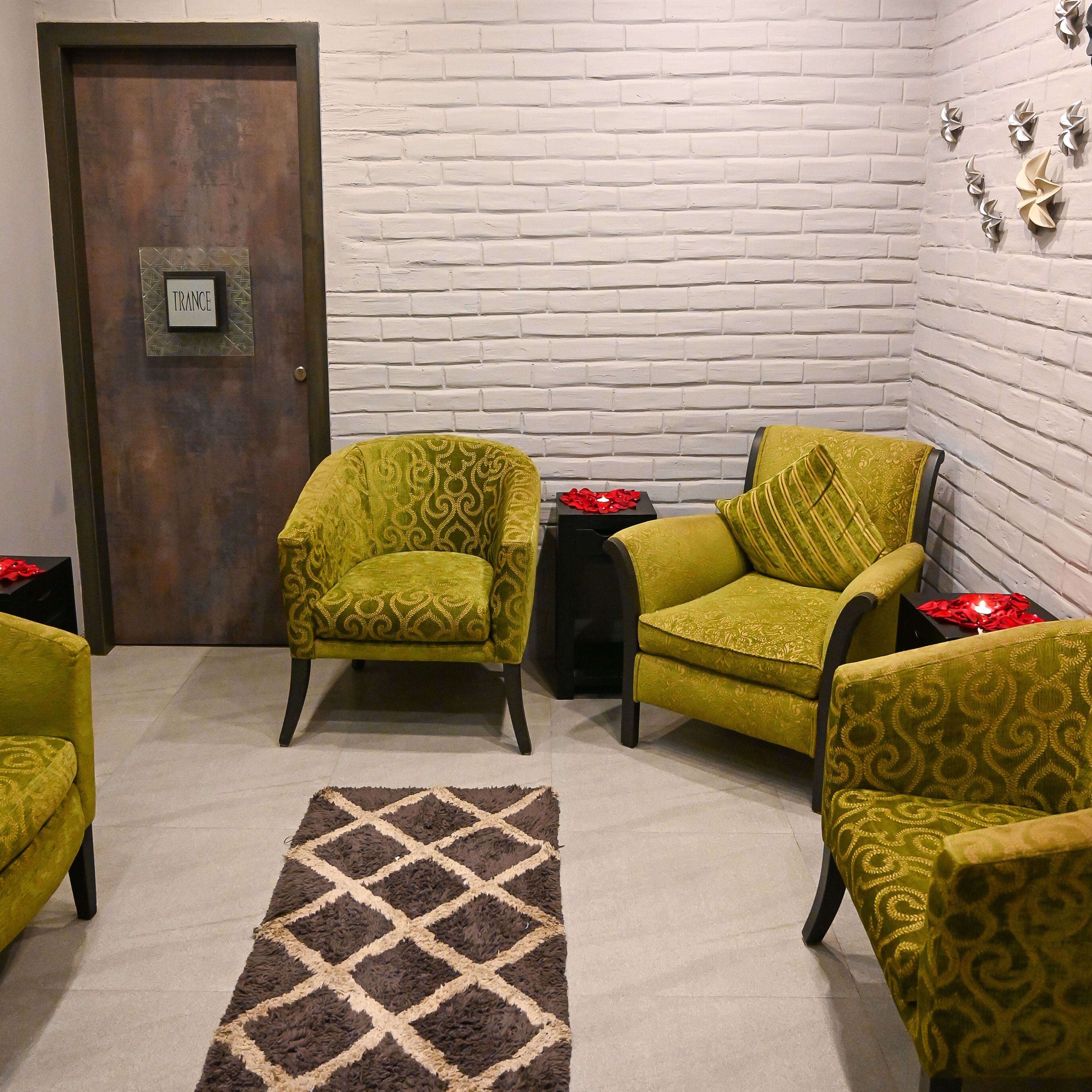 Una habitación con pared de ladrillo blanco, dos sillones verdes con estampado, una mesita auxiliar negra con una vela roja y una alfombra marrón y beige.