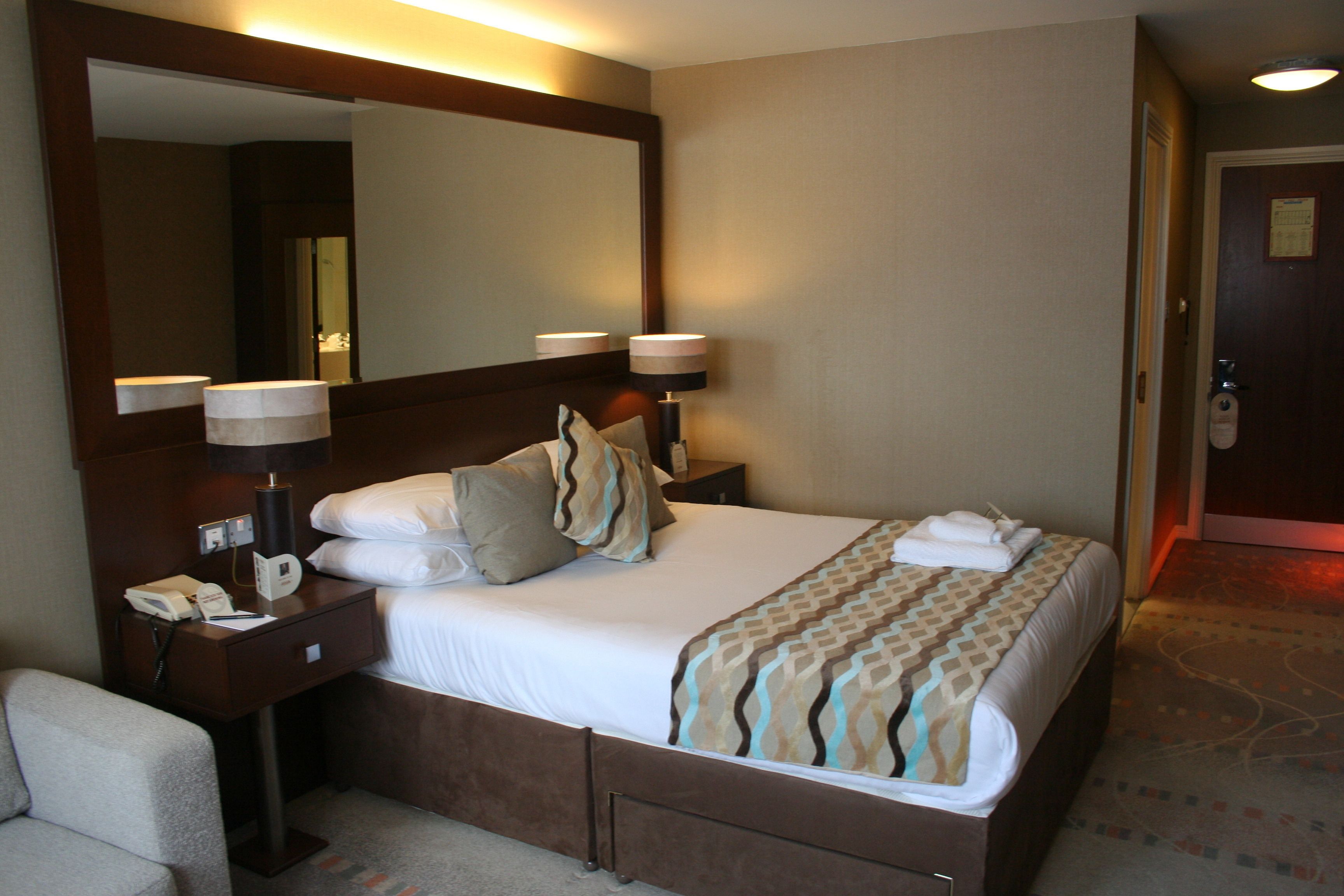 Habitación de hotel con una cama grande, una mesita de noche con lámpara, un espejo y una silla.