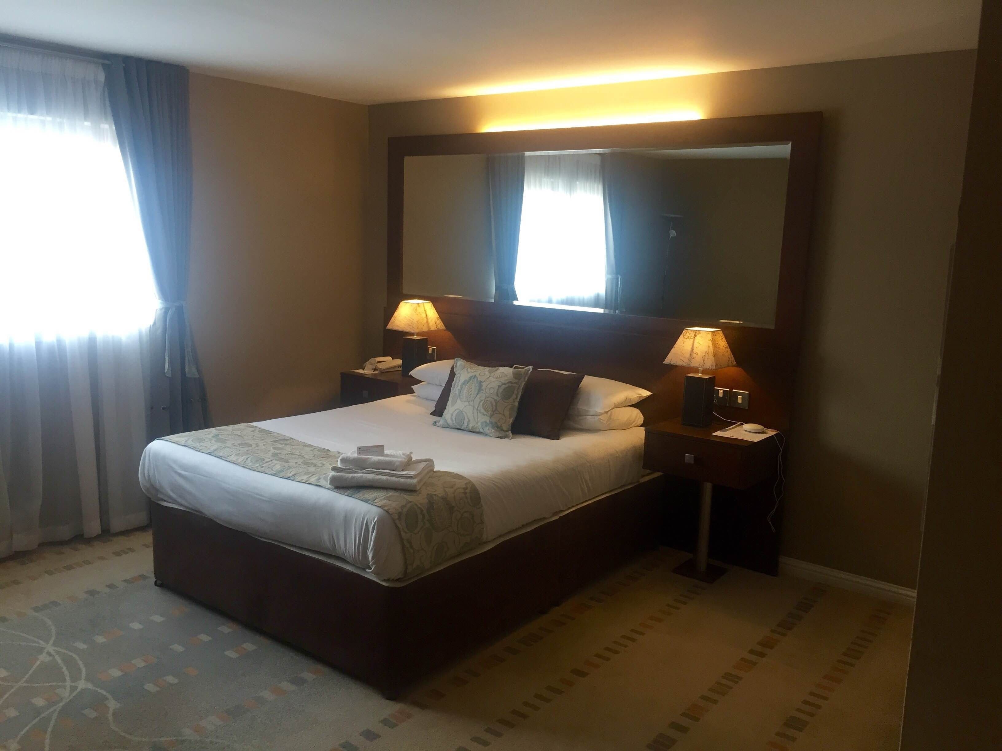 Ein Hotelzimmer mit einem großen Bett, zwei Nachttischen mit Lampen, einem großen Spiegel über dem Bett und einem Fenster mit Vorhängen.