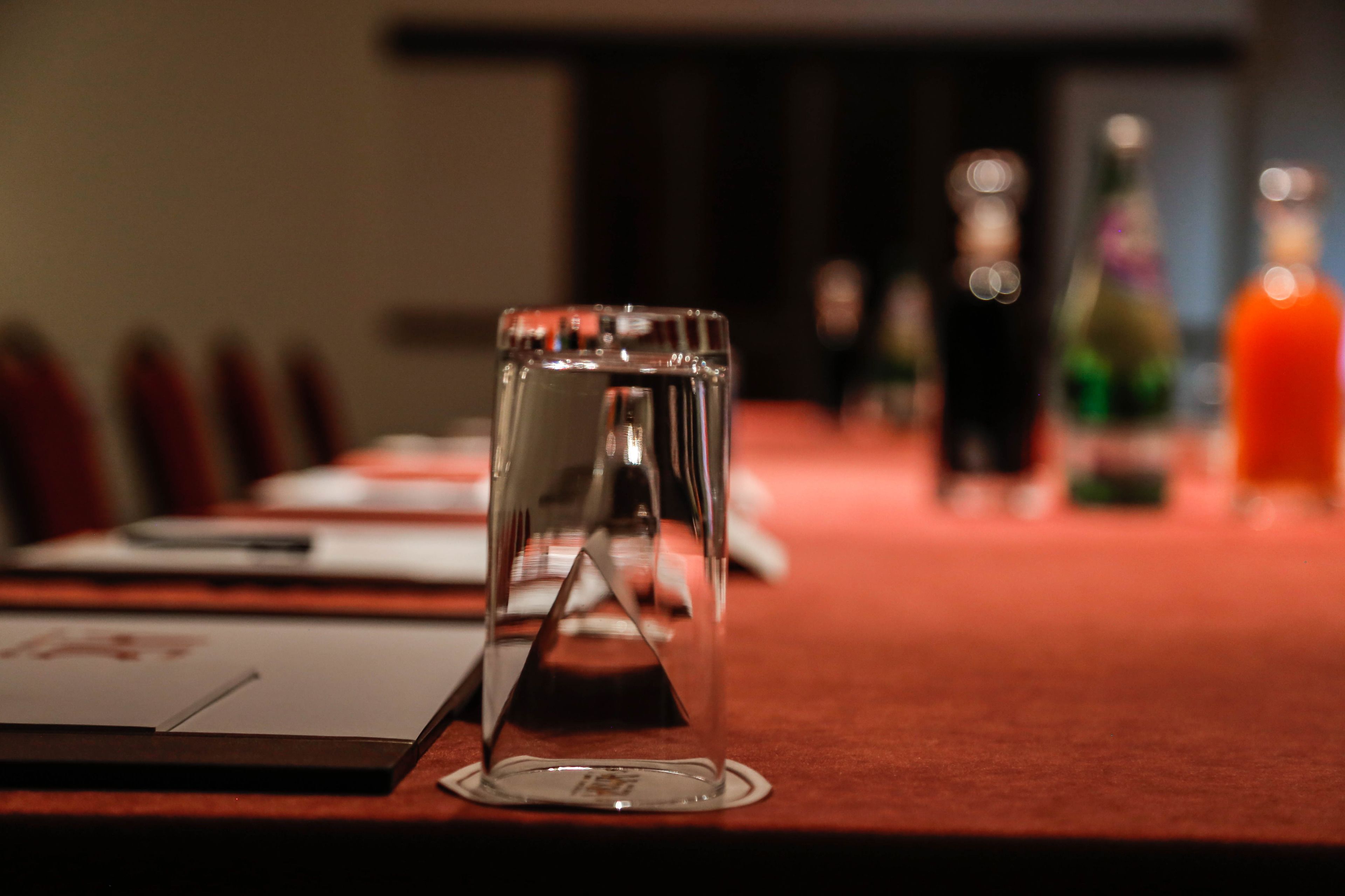 A conference table with a clear glass centerpiece, documents, and bottles in the background.
