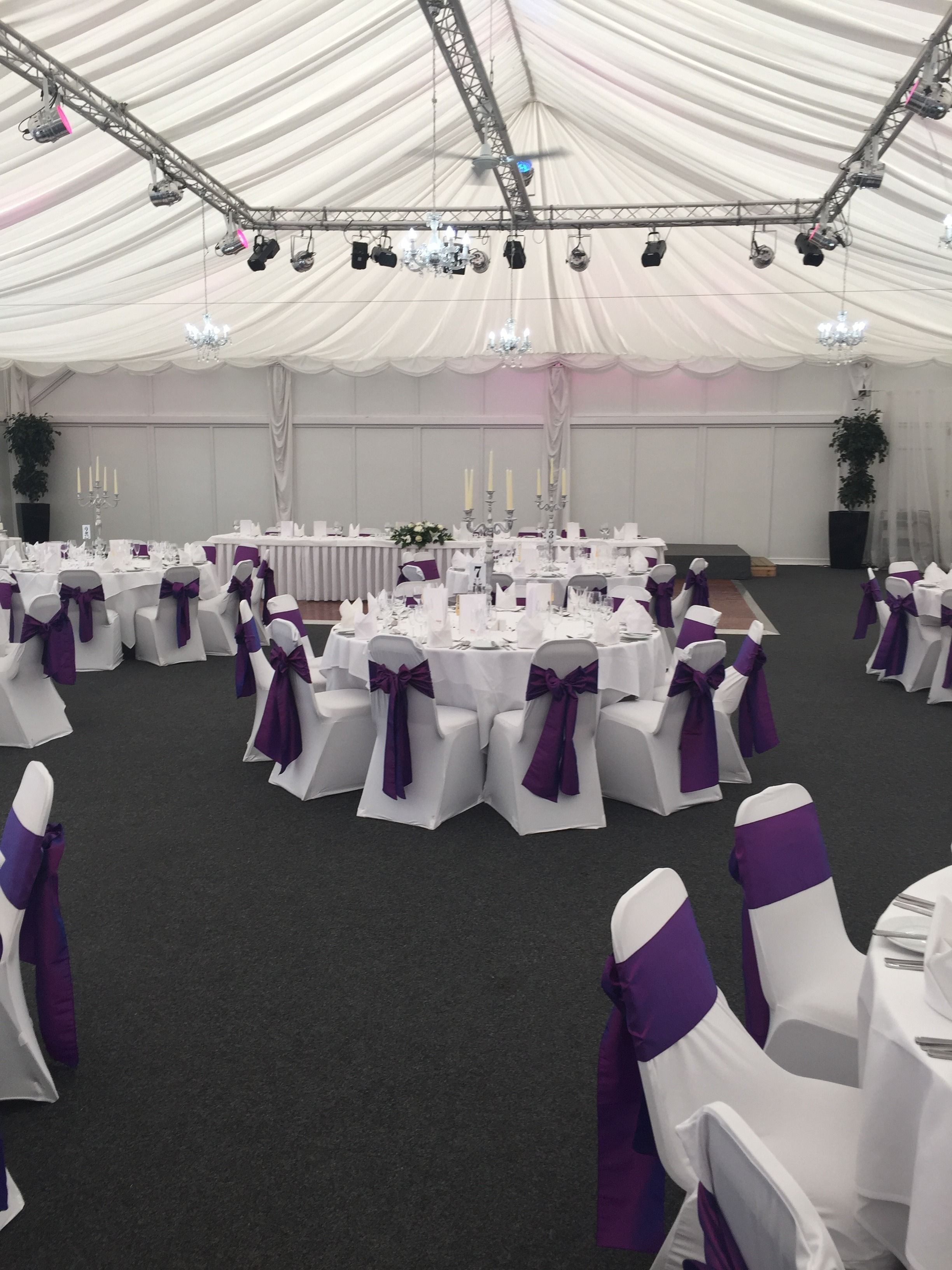 A tented event space with round tables, white chairs with purple sashes, and a stage with a candelabra.