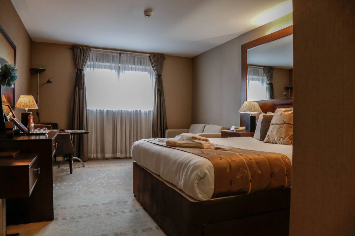 A hotel room with a bed, a desk, a chair, a lamp, and a window with curtains.