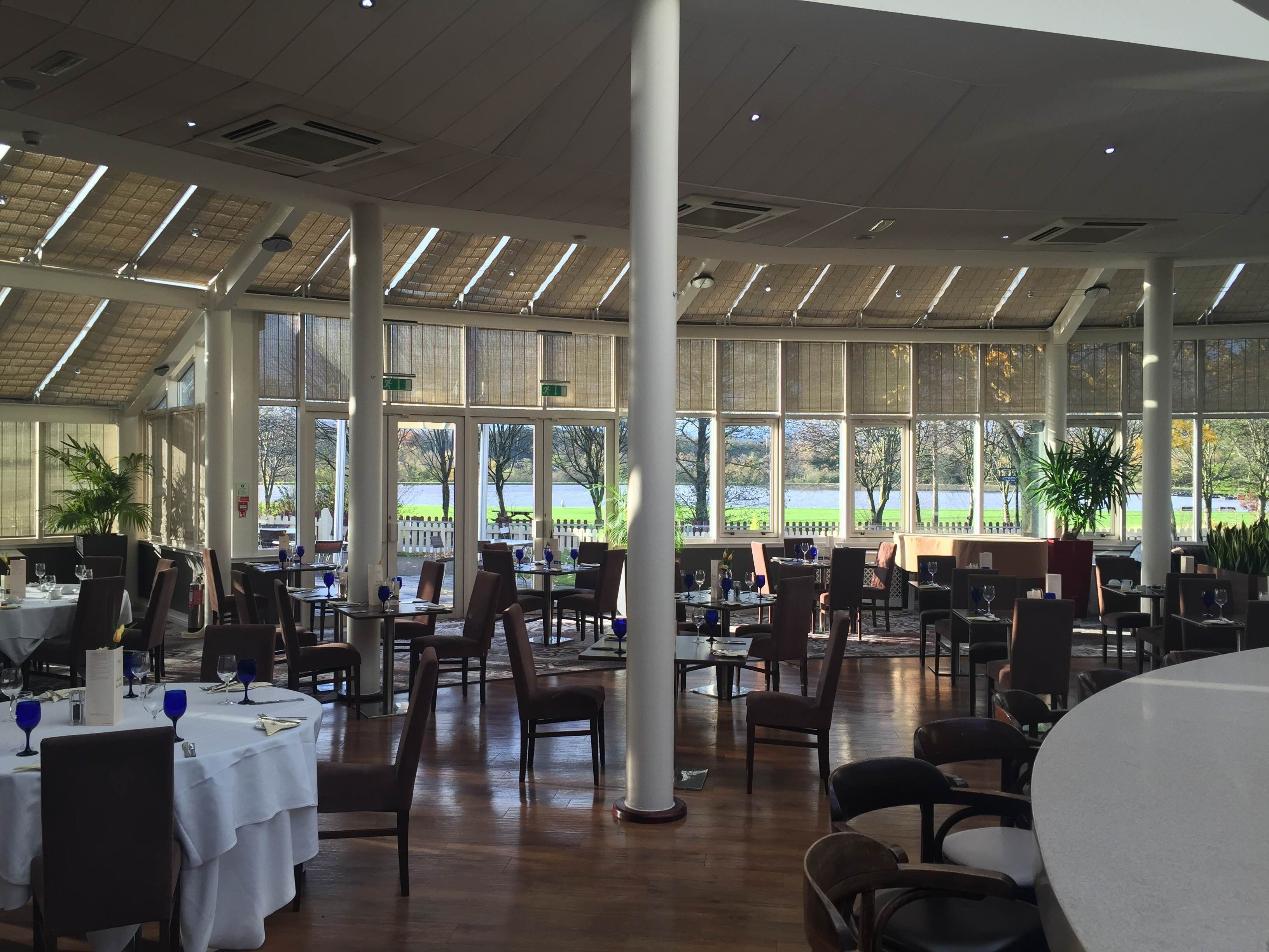 A spacious indoor dining area with high ceilings, large windows, and a view of a lake. Tables are set with white tablecloths and blue wine glasses.