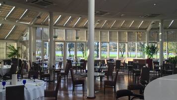 A spacious indoor dining area with high ceilings, large windows, and a view of a lake. Tables are set with white tablecloths and blue wine glasses.