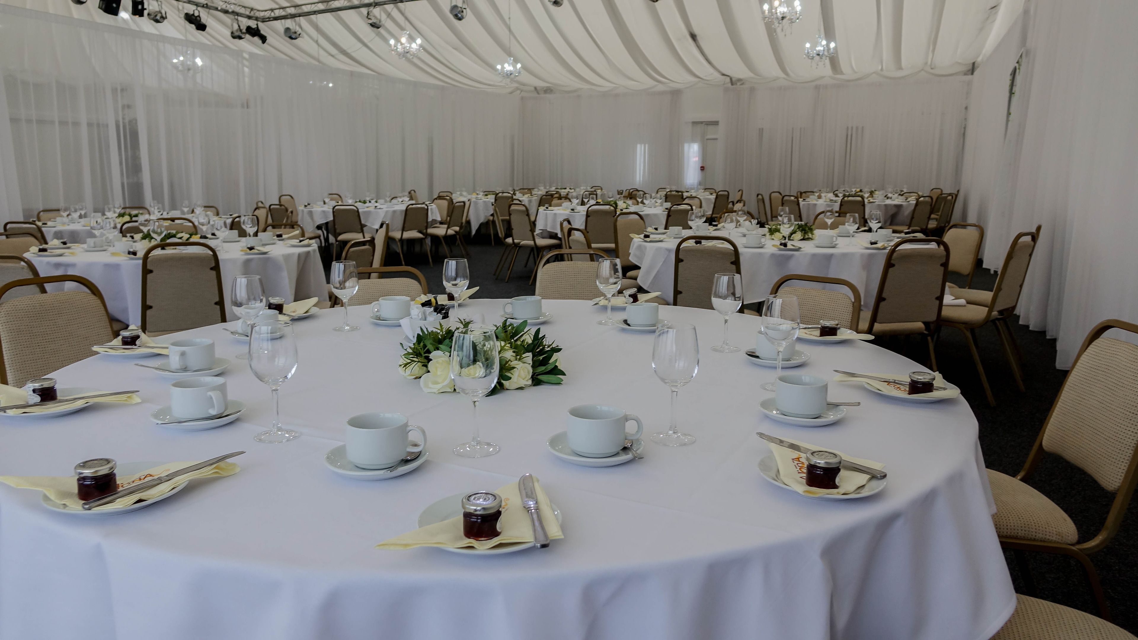 Een feestzaal met ronde tafels gedekt voor een formeel evenement, met witte tafelkleden, glazen en bloemstukken als decoratie.