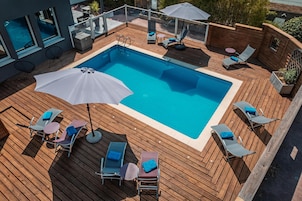 A rectangular swimming pool with a wooden deck surrounding it, equipped with lounge chairs and umbrellas.