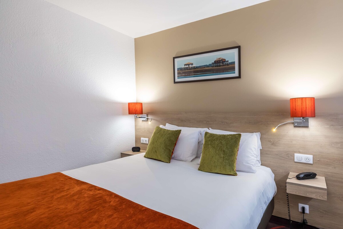 Una habitación de hotel con una cama, dos mesitas de noche, una lámpara, un teléfono y un cuadro en la pared.
