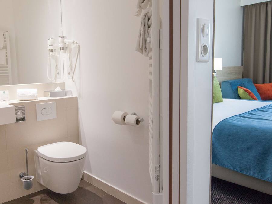 A hotel room with a bathroom, a bed with colorful pillows, a white toilet, and a mirror.