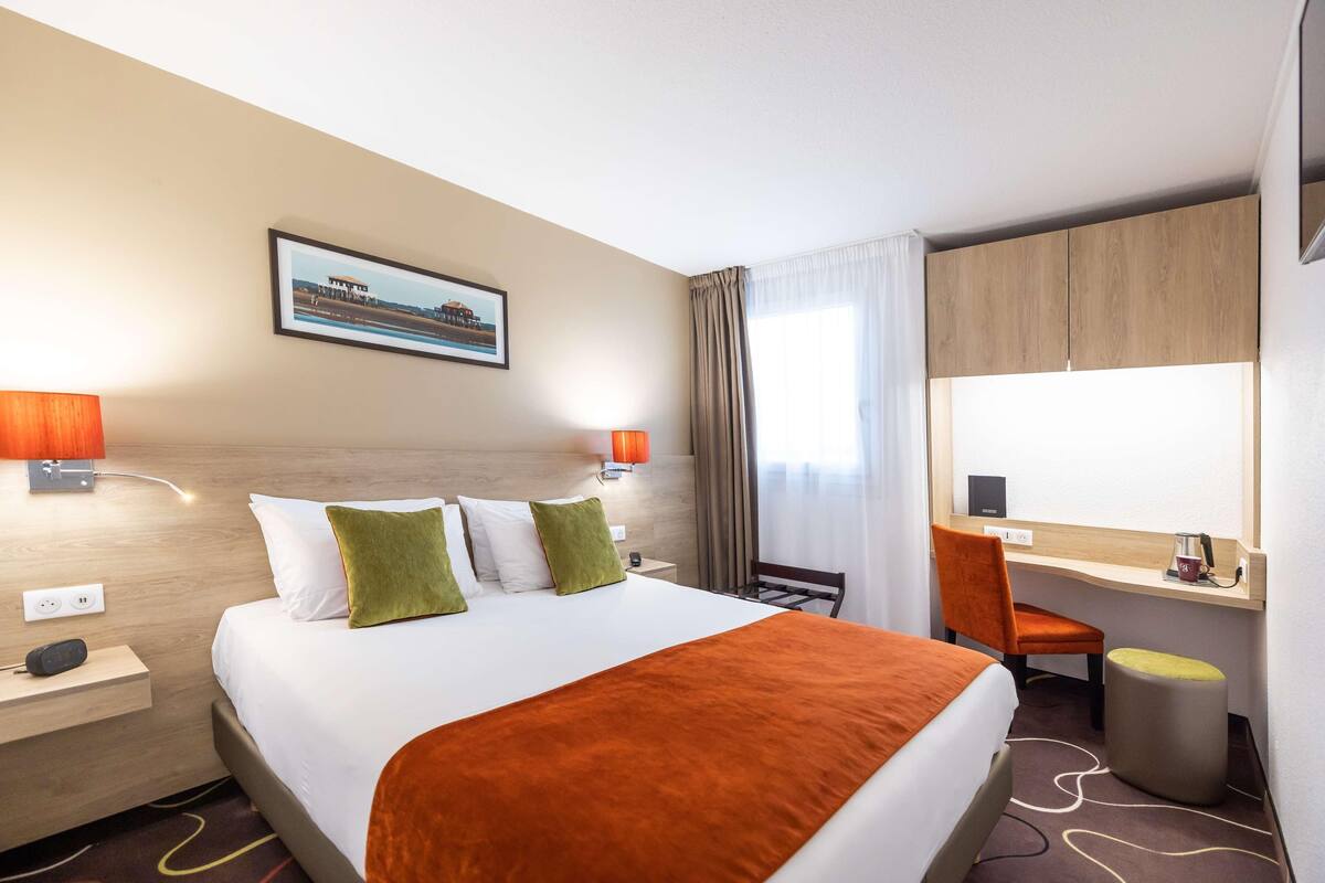 A hotel room with a bed, two bedside lamps, a desk with a chair, a small stool, a mirror, and a framed picture on the wall.