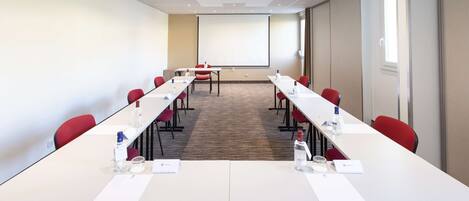 Un salón de conferencias con una larga mesa, sillas rojas, una pantalla de proyector y piso alfombrado.