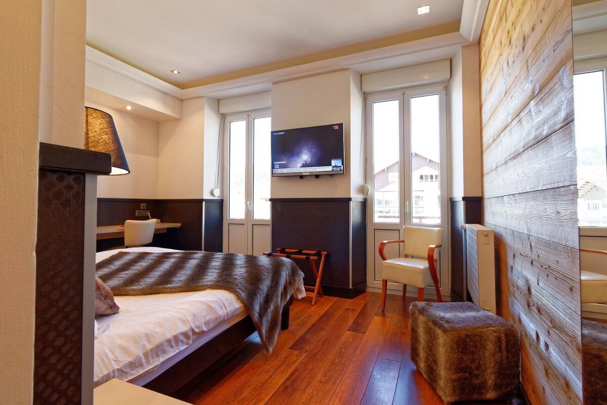 Habitación de hotel con cama, una silla, televisor montado en la pared y ventana con vistas a edificios.