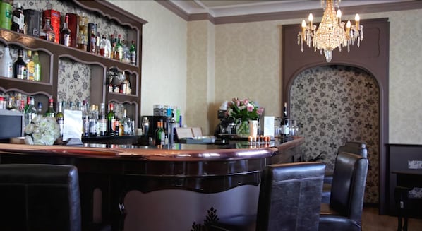 A bar area with a counter, shelves of liquor bottles, a chandelier, and a floral wallpapered wall. - Hotel des Bains (Gerardmer)