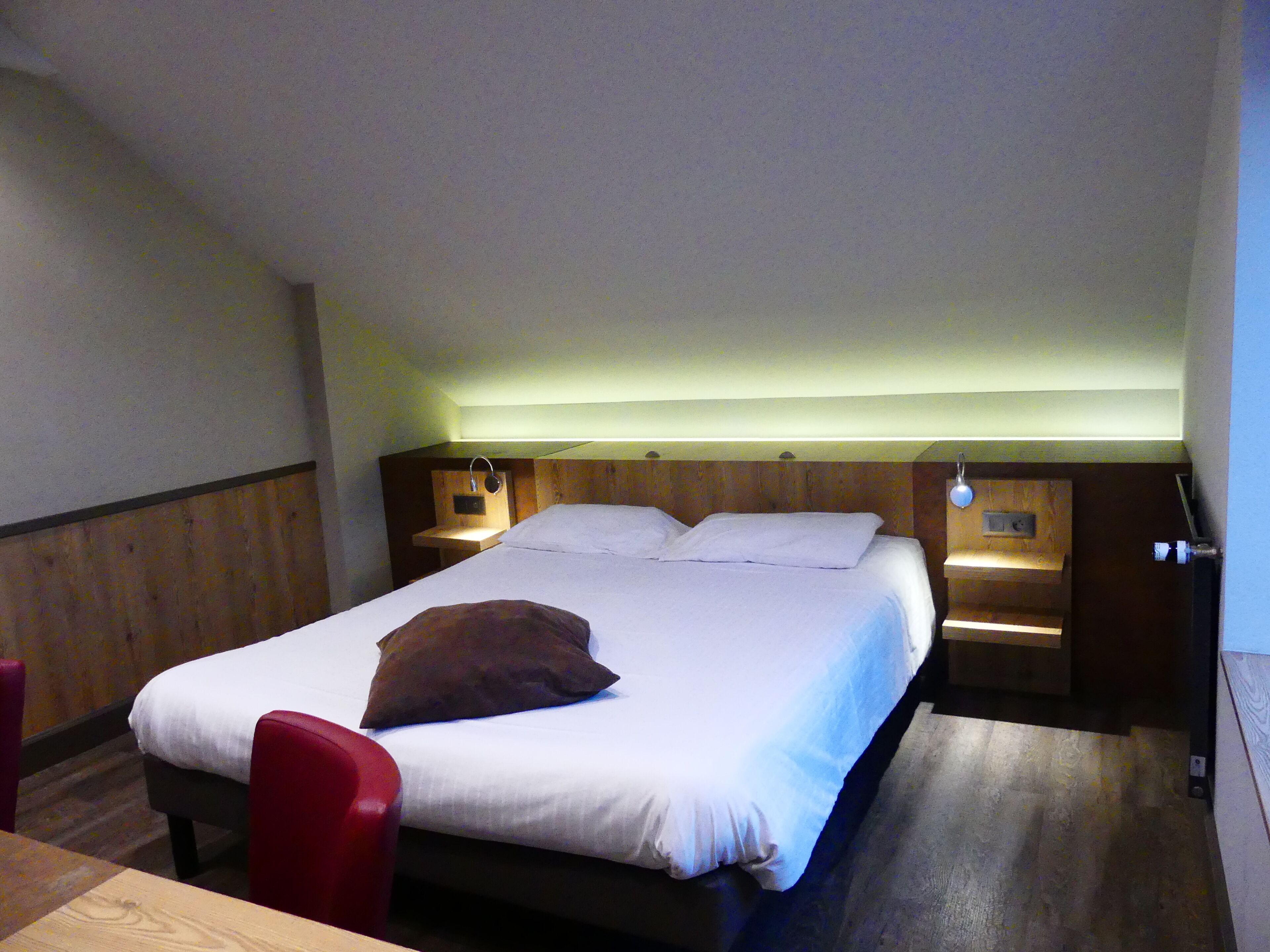 A single bed with a wooden headboard, two bedside tables with lamps, a red chair, and a wooden floor.