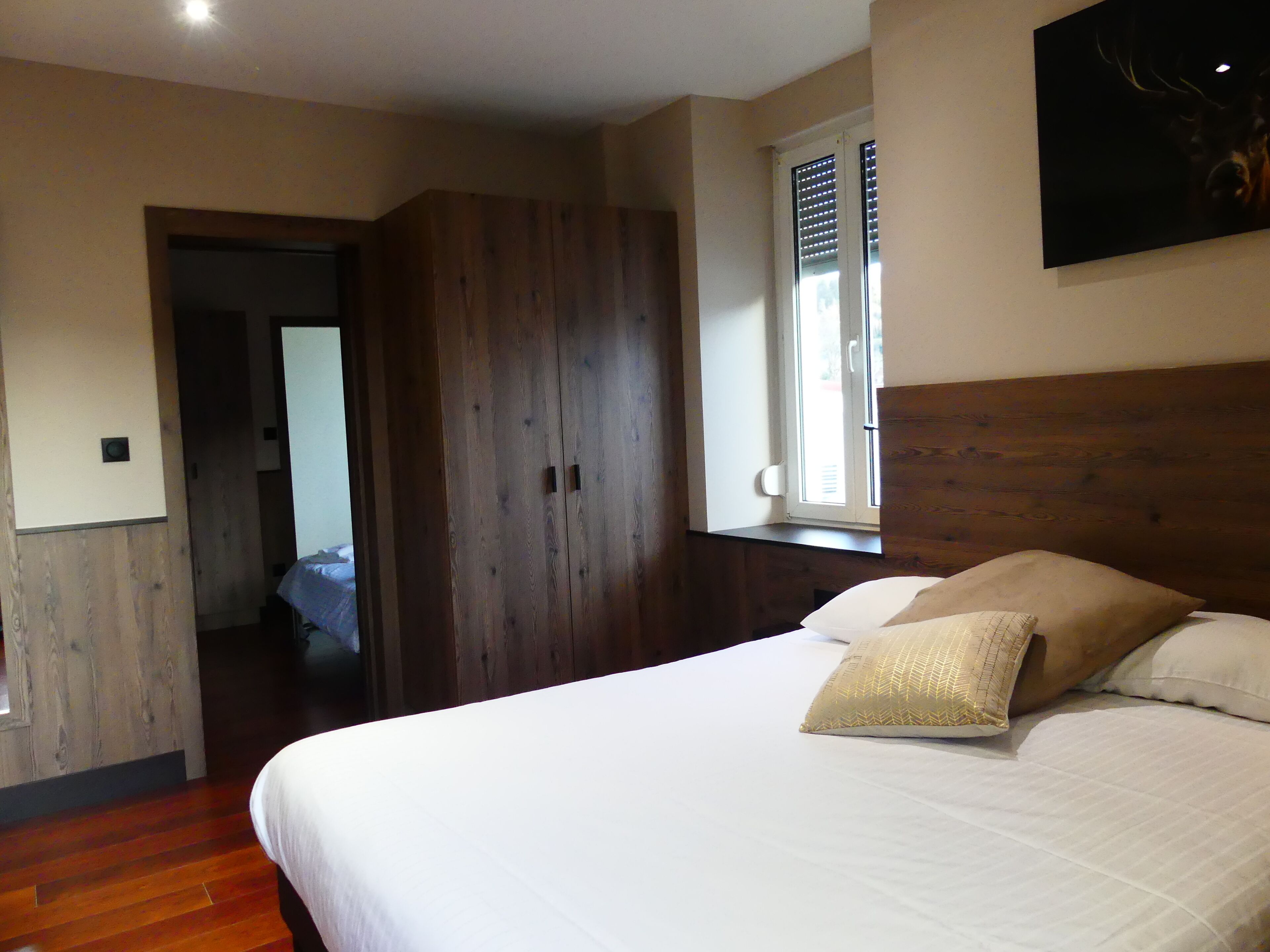 Une chambre avec une armoire en bois, un lit avec une literie blanche et une fenêtre avec des stores.