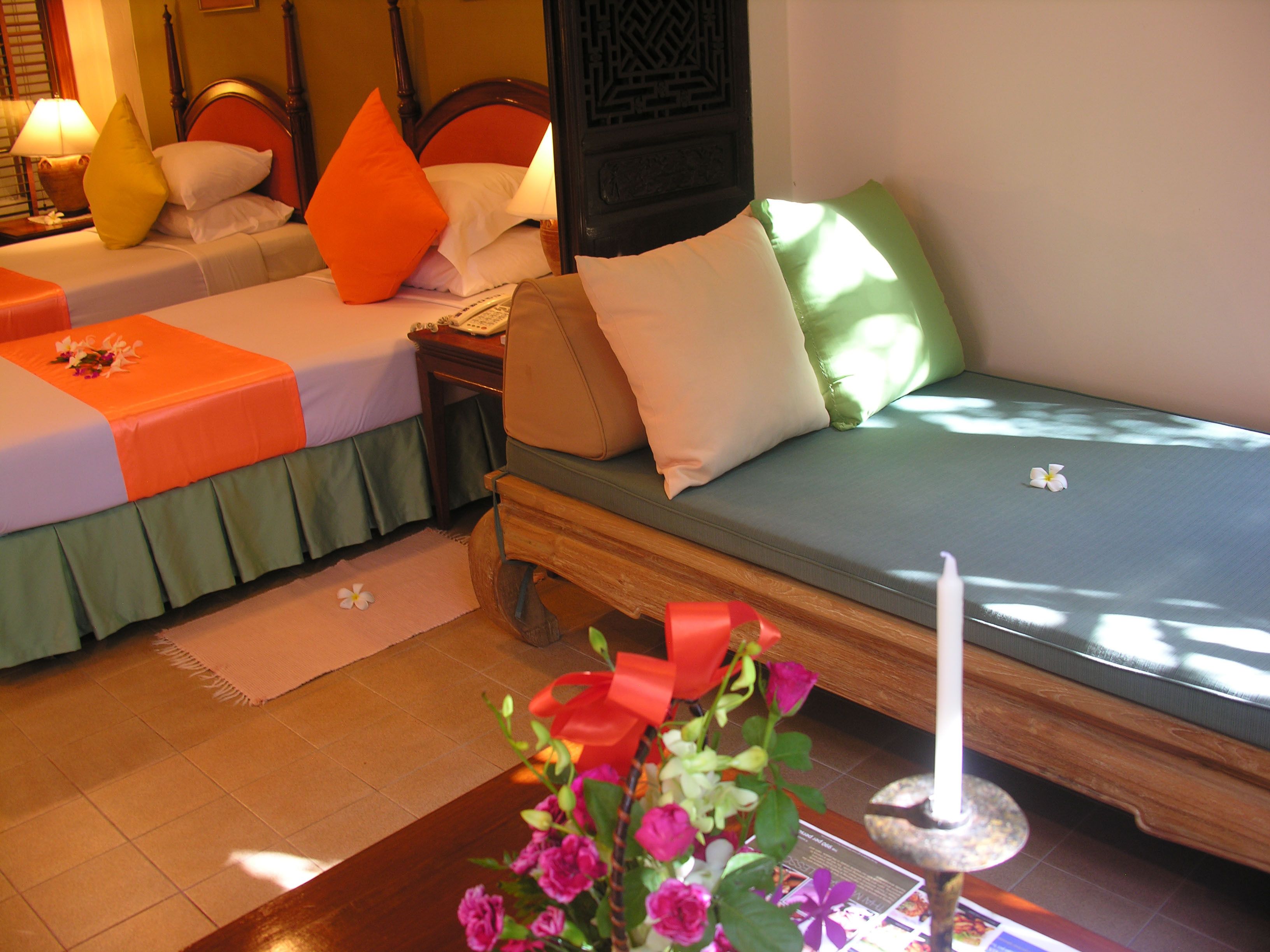A hotel room with two beds, each with a different color bedspread and pillows. There is a nightstand with a lamp and a vase of flowers in the foreground.