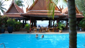 A pool area with two people swimming, surrounded by wooden structures with a tiled roof, and potted plants. - White House Beach Resort (Koh Samui)