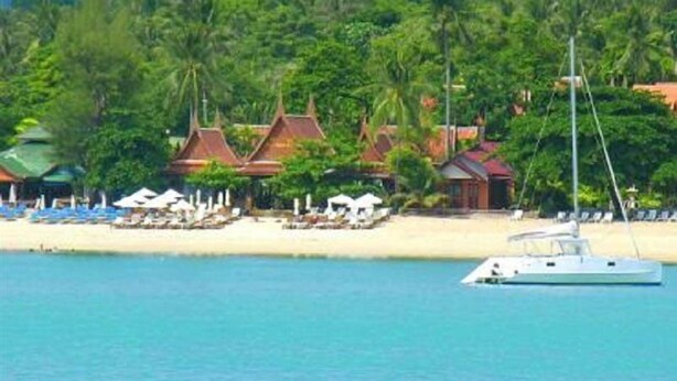 A beachfront hotel with a boat docked near the shore, surrounded by lush greenery and traditional architecture. - White House Beach Resort (Koh Samui)