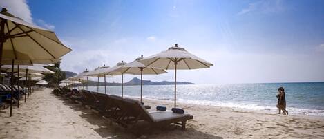A beach with rows of sun loungers and umbrellas, a clear sky, and the ocean in the background.