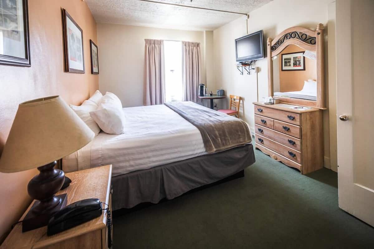 A hotel room with a bed, bedside lamp, telephone, dresser, mirror, television, and curtains.