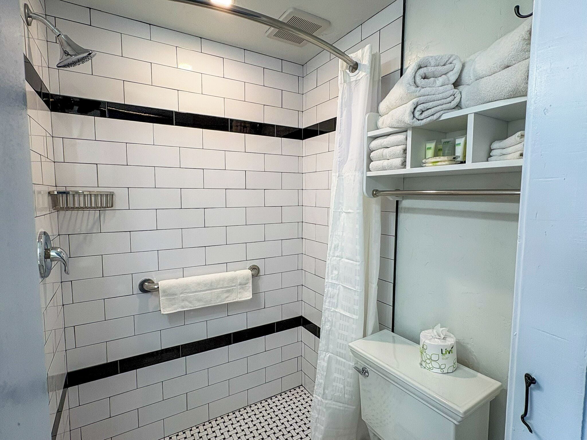 A modern bathroom with white subway tiles, a black accent strip, a glass shower enclosure, a toilet with a Livi toilet paper dispenser, and a towel rack with folded towels.