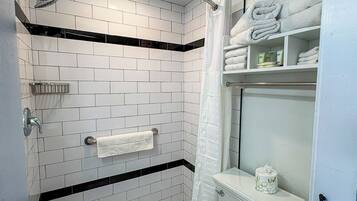 A modern bathroom with white subway tiles, a black accent strip, a glass shower enclosure, a toilet with a Livi toilet paper dispenser, and a towel rack with folded towels.