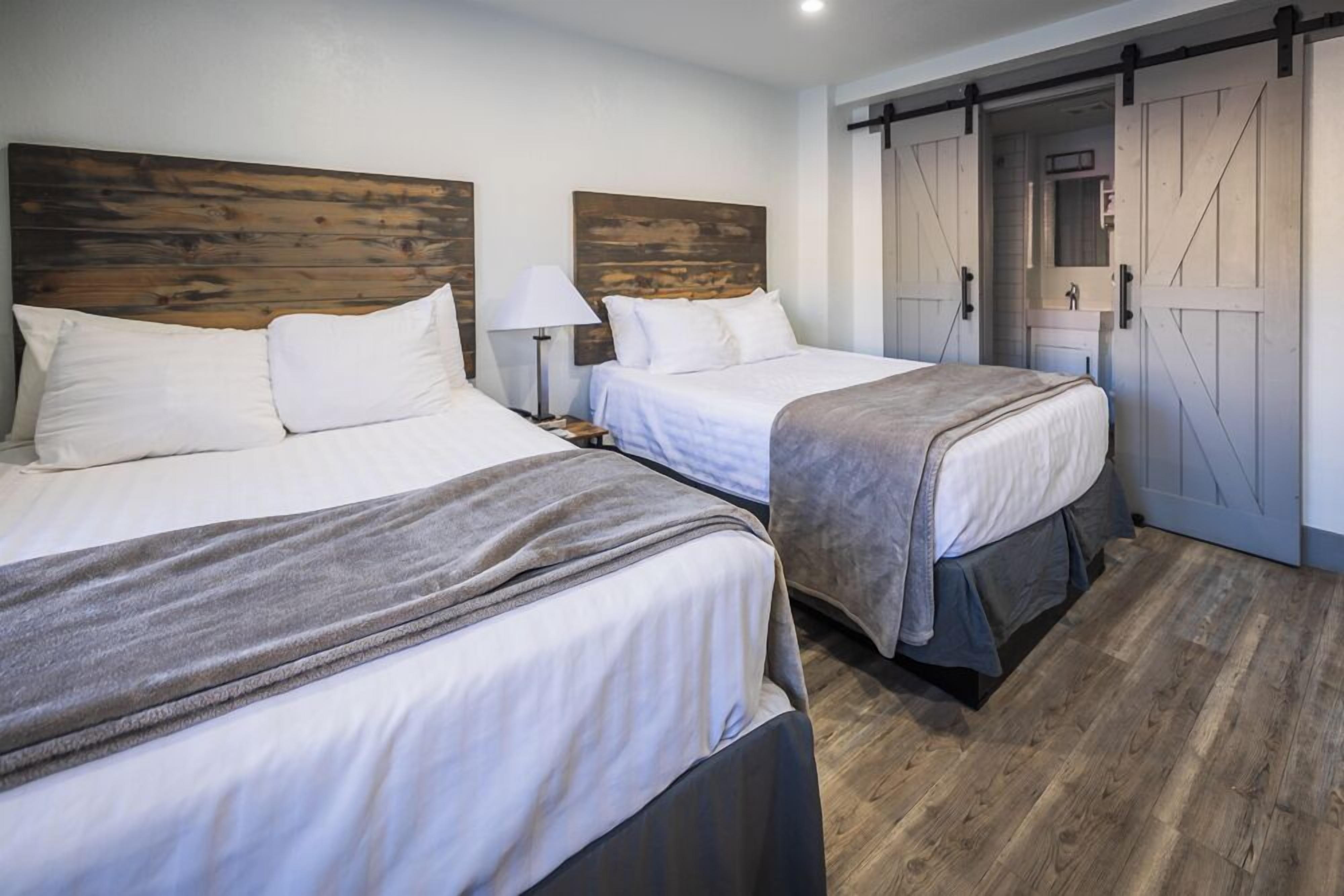 Quarto de hotel com duas camas, cabeceiras de madeira, uma porta corrediça estilo celeiro e piso de madeira.
