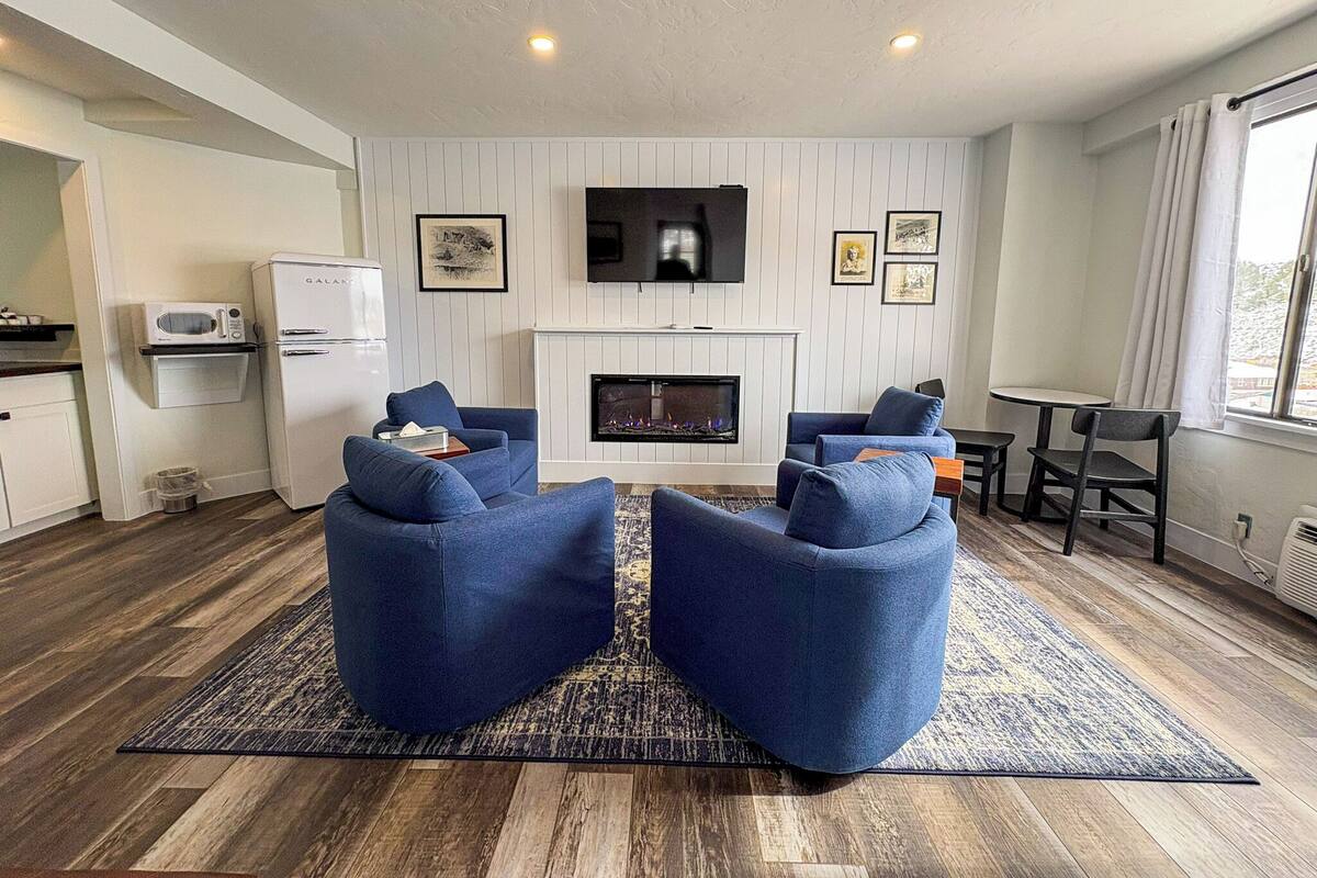 A modern living room with a fireplace, blue sofas, a flat-screen TV, and a kitchen area with a microwave and refrigerator.