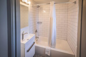 Un baño moderno con paredes de azulejos blancos, una bañera blanca, una ducha con puerta de cristal, un lavabo blanco con espejo y una toalla colgando de la bañera.