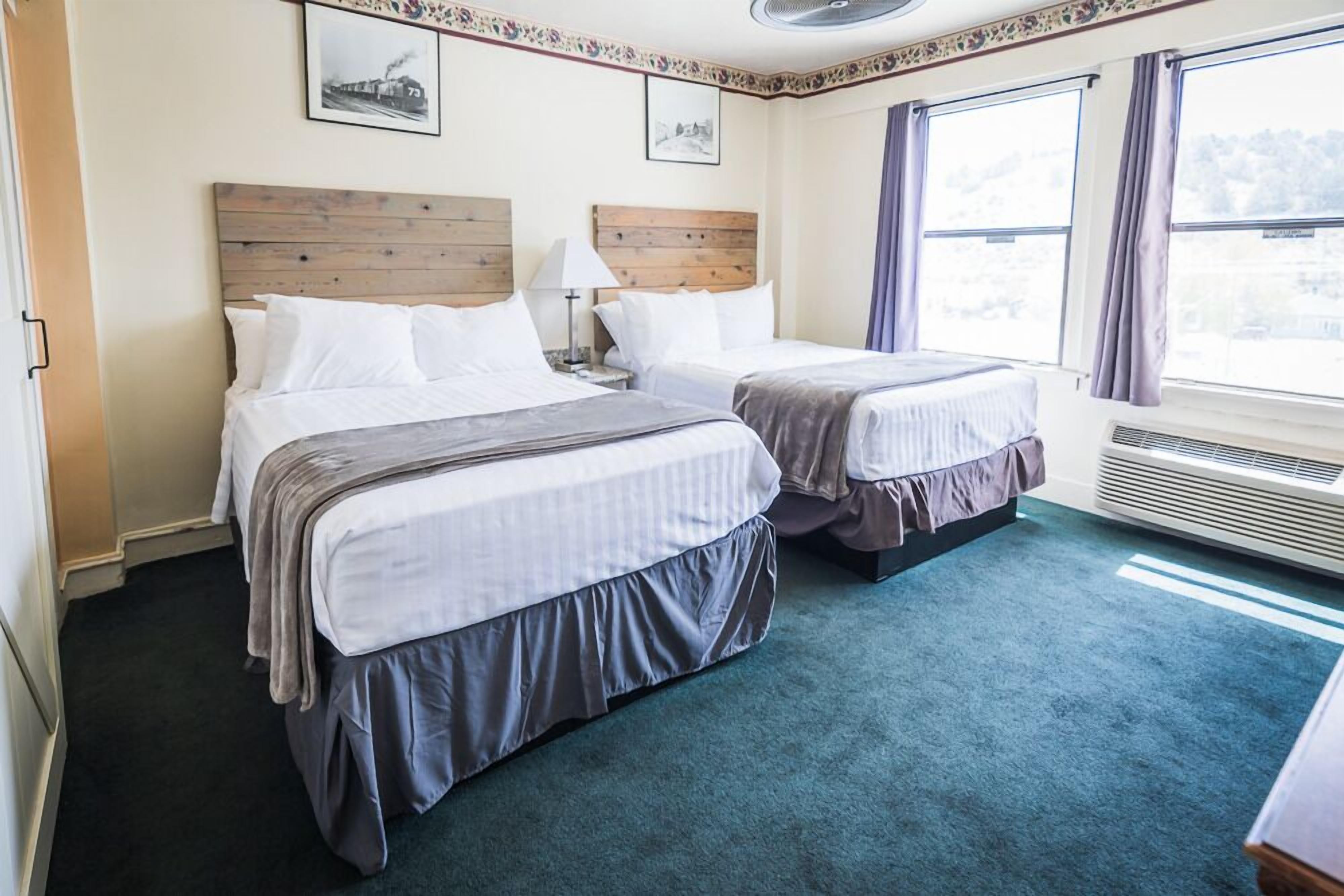 Une chambre d’hôtel avec deux lits, une tête de lit en bois, une grande fenêtre avec des rideaux et un sol recouvert de moquette.