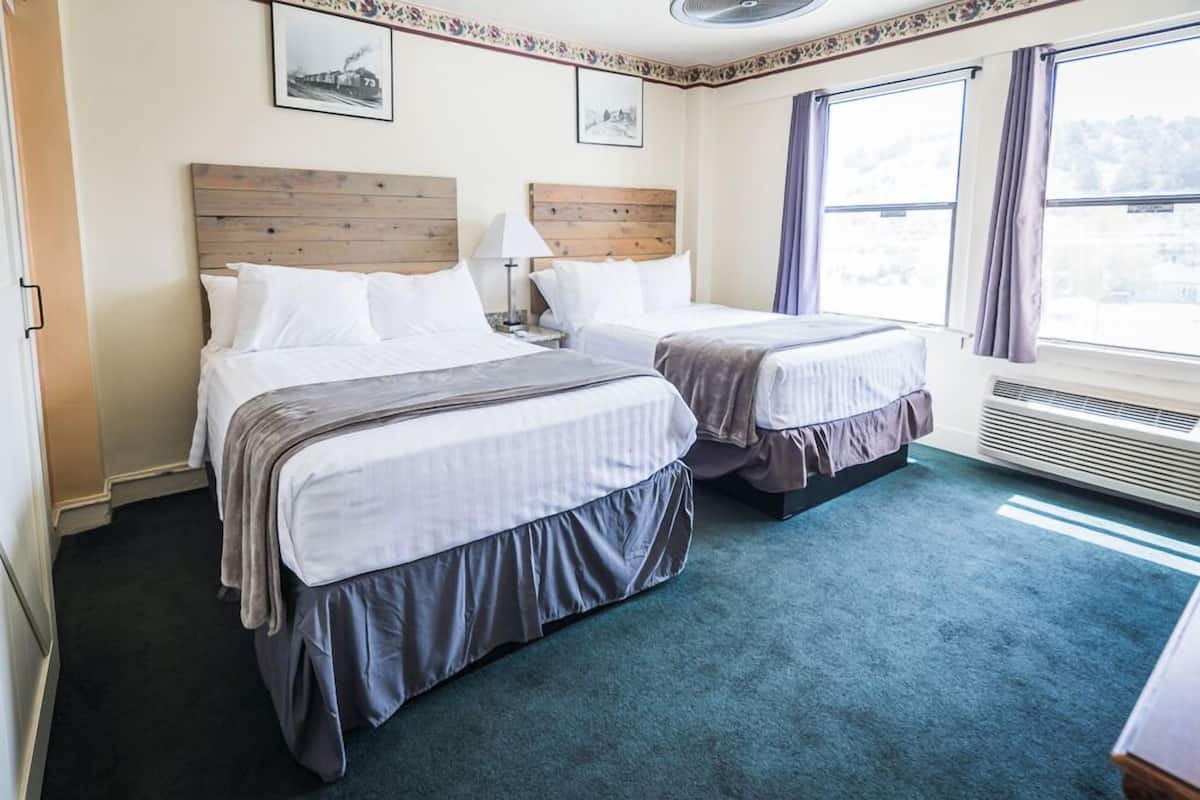 Habitación de hotel con dos camas, cabecero de madera, ventana grande con cortinas y suelo alfombrado.