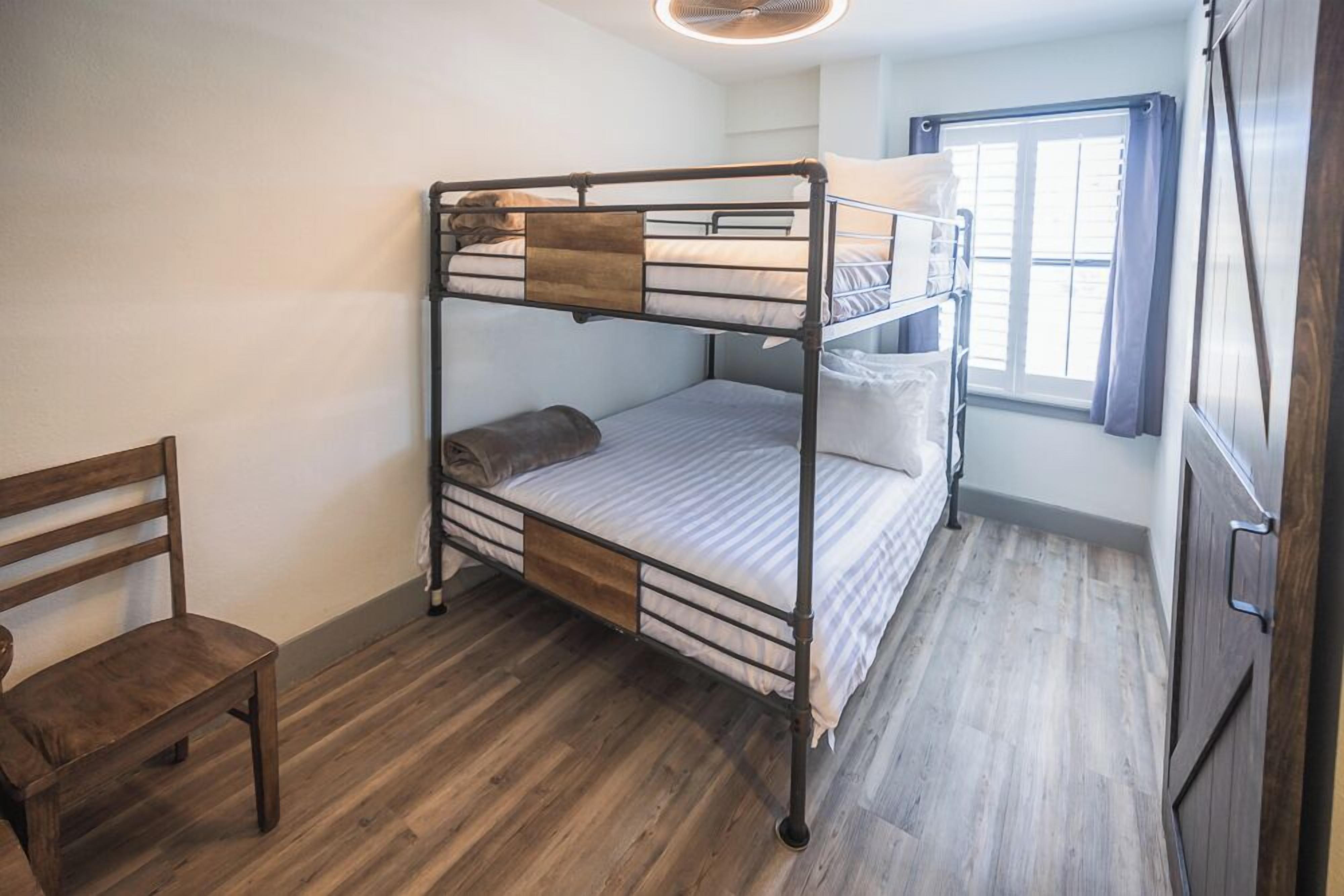 Una stanza con un letto a castello, una panca di legno, una finestra con tende e una porta.