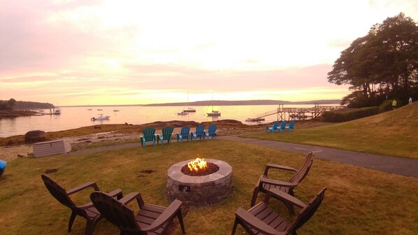 A fire pit with chairs, a grassy area, and a view of boats on the water at sunset. - Smuggler's Cove Inn (East Boothbay)