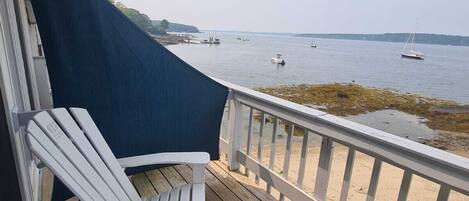 Une terrasse en bois avec un fauteuil Adirondack blanc, une balustrade blanche et une vue sur l’eau, où l’on peut apercevoir des bateaux.