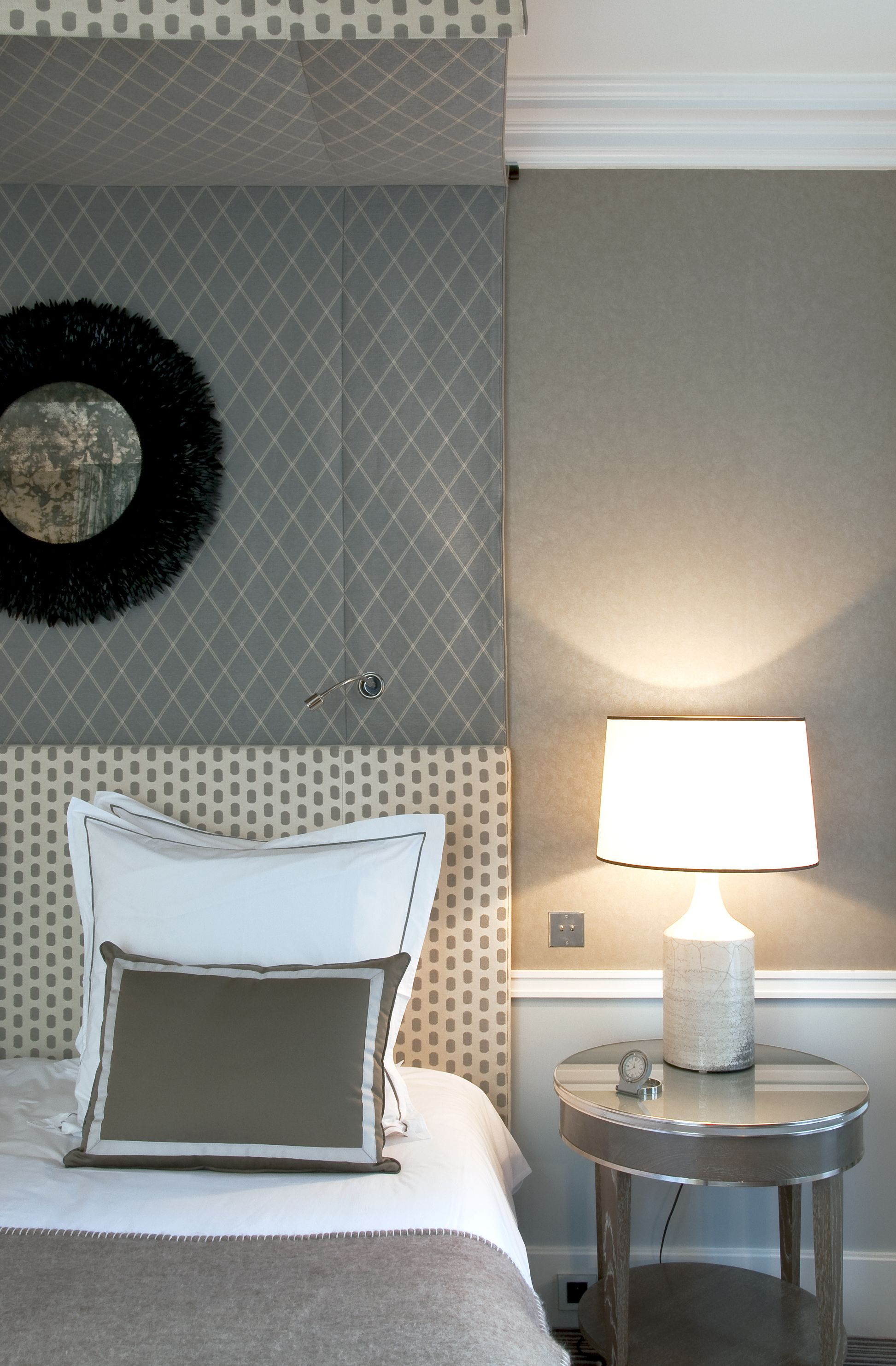 A neatly made bed with two pillows, a round wall clock, and a table lamp on a bedside table.