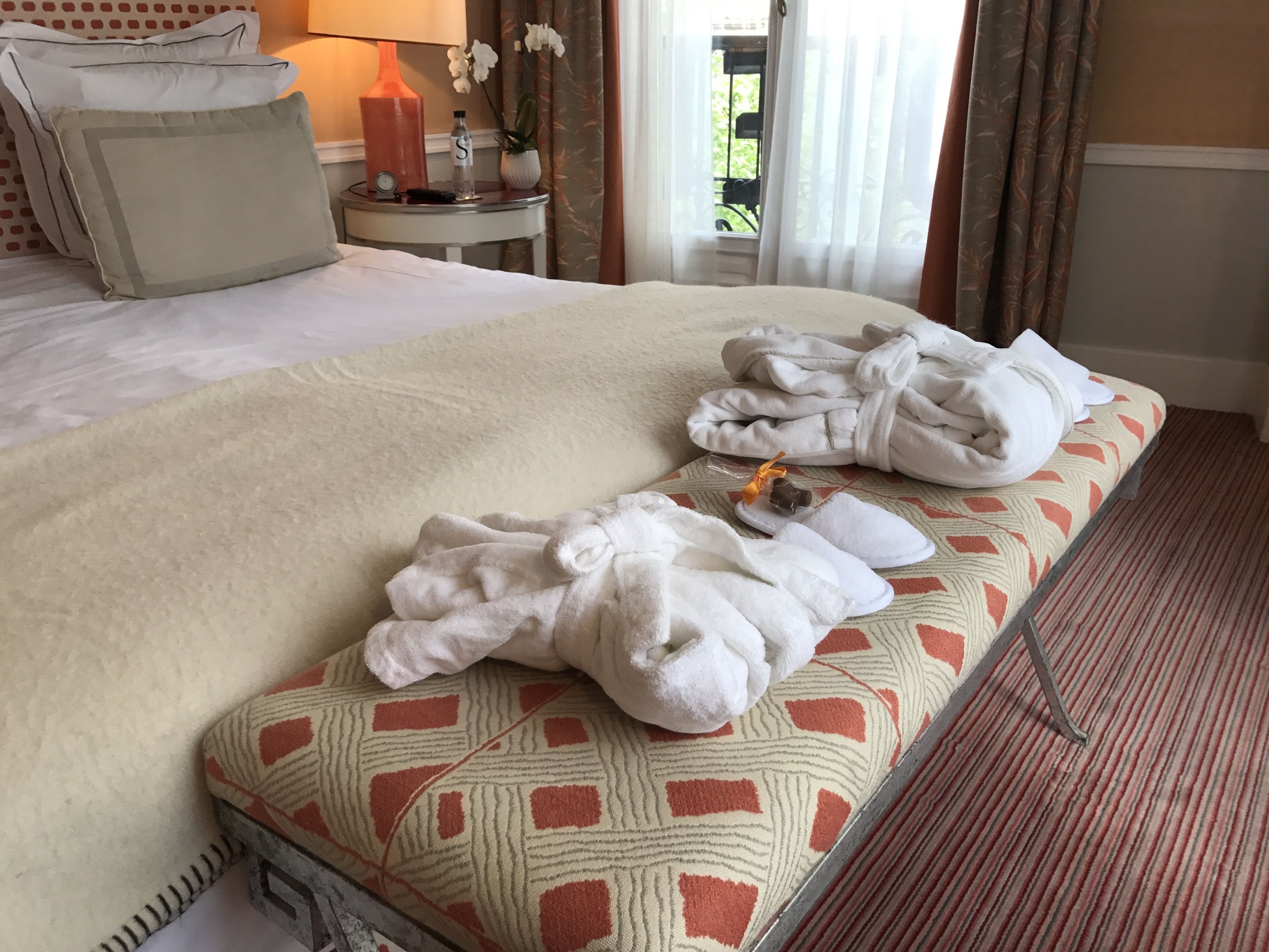 Een hotelkamer met een bed, nachtkastje met lamp, vaas met bloemen en een raam met gordijnen.