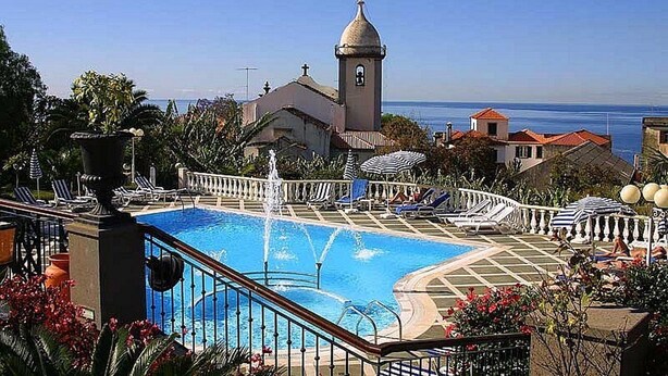 Una piscina con una fuente, rodeada de sillas de descanso y sombrillas, con una iglesia y vista al océano de fondo.