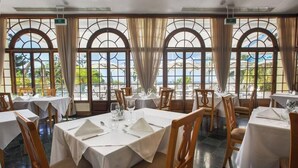 Un restaurante con mesas dispuestas para comer, ventanas amplias con cortinas y vistas a zonas verdes.