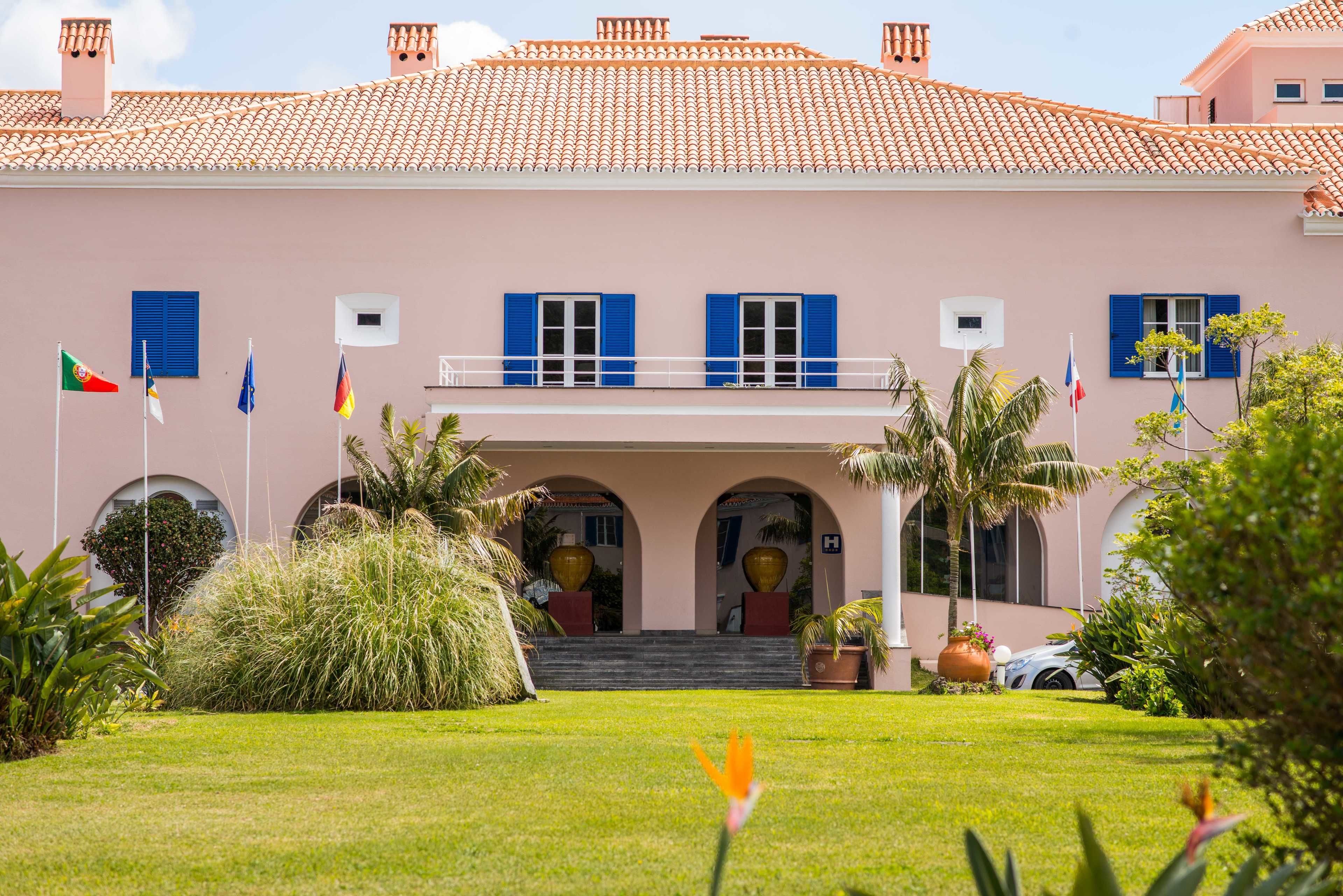 Un edificio rosa con contraventanas azules, un porche cubierto y varias banderas.