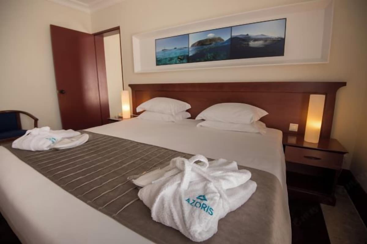 A hotel room with a large bed, two bedside tables, a painting above the bed, and a door to the left.