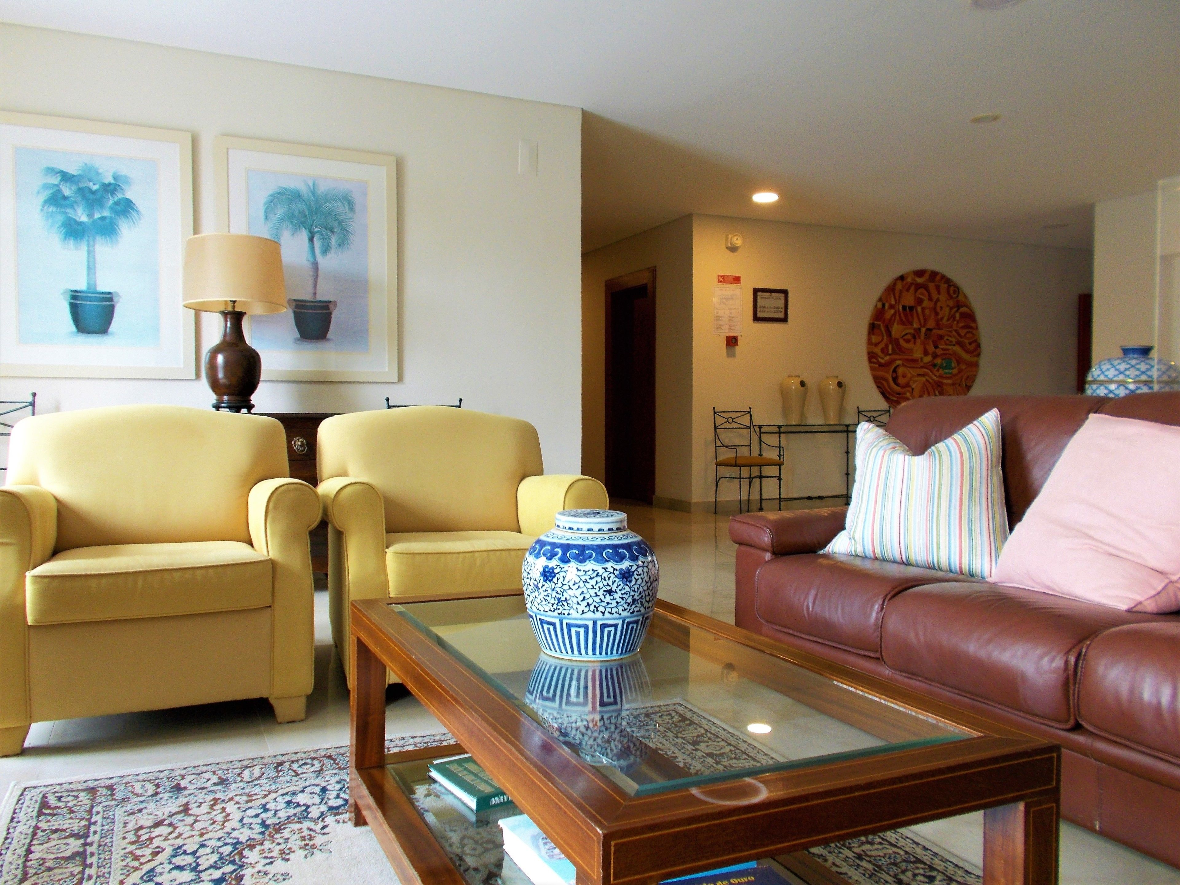 A living room with two yellow armchairs, a brown leather sofa, a glass coffee table with a blue and white vase, and a patterned rug.