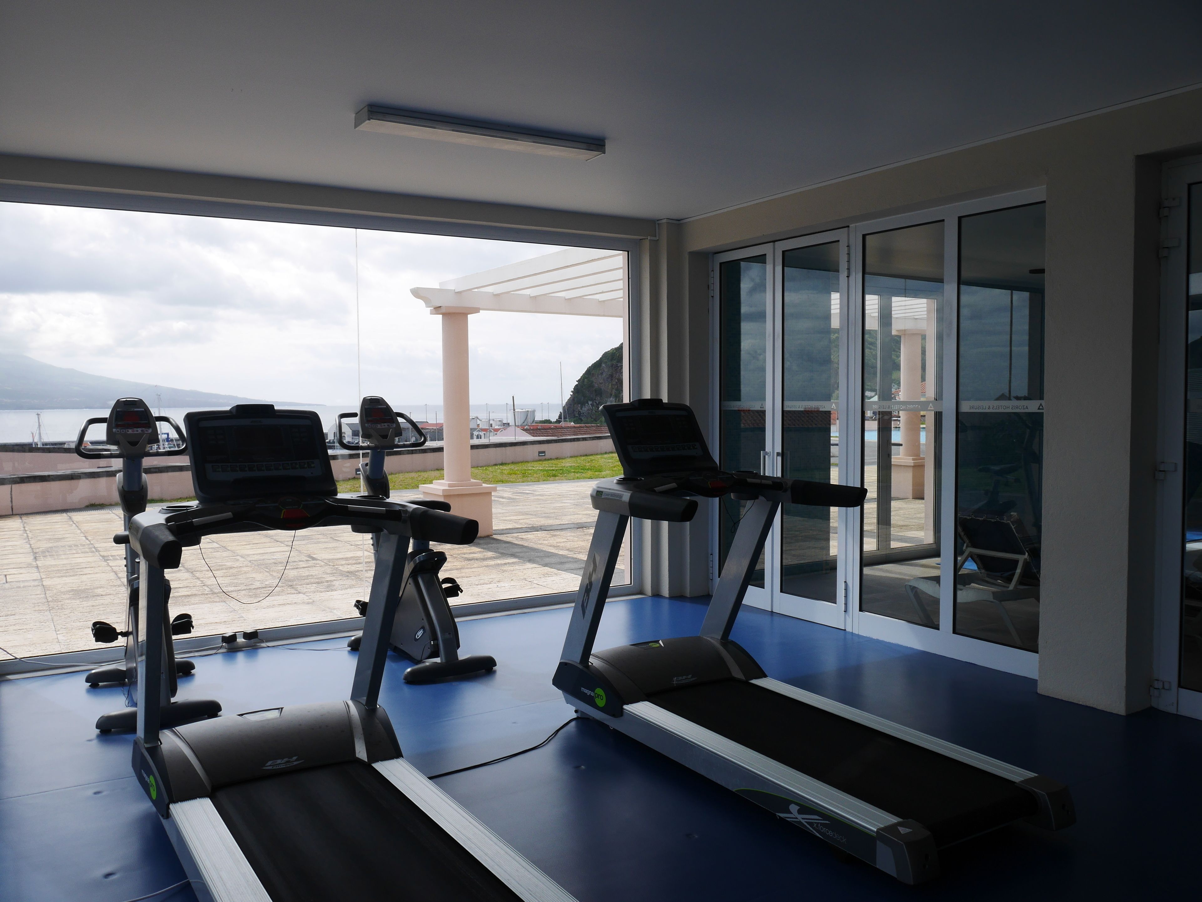 A gym with treadmills and exercise bikes, large windows with a view of the sea and mountains, and a covered outdoor area.