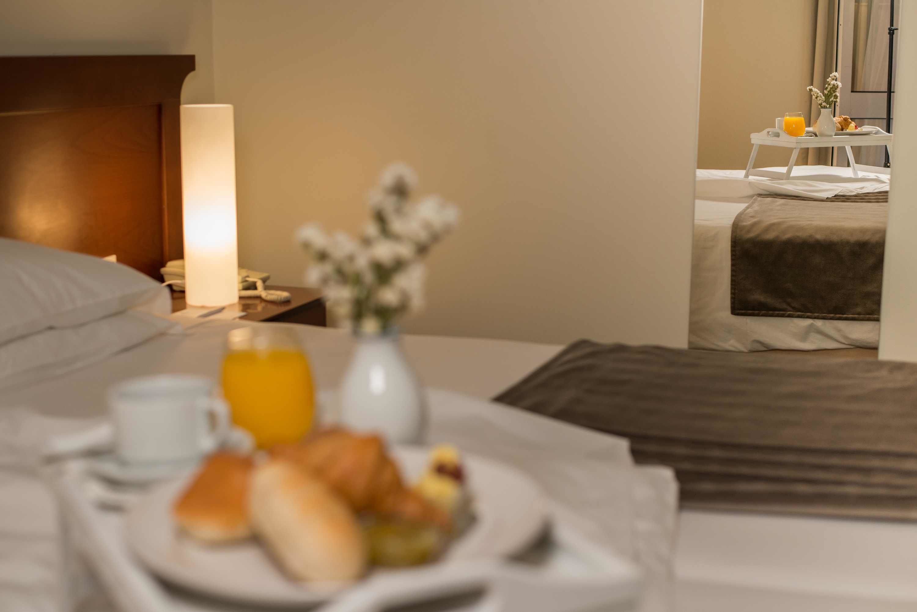 A hotel room with a neatly made bed, a bedside table with a lamp, a tray with breakfast items, and a small table with a vase of flowers.