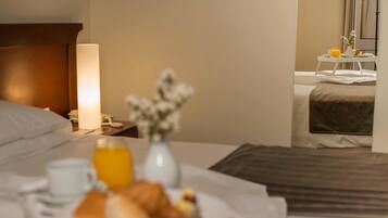 A hotel room with a neatly made bed, a bedside table with a lamp, a tray with breakfast items, and a small table with a vase of flowers.