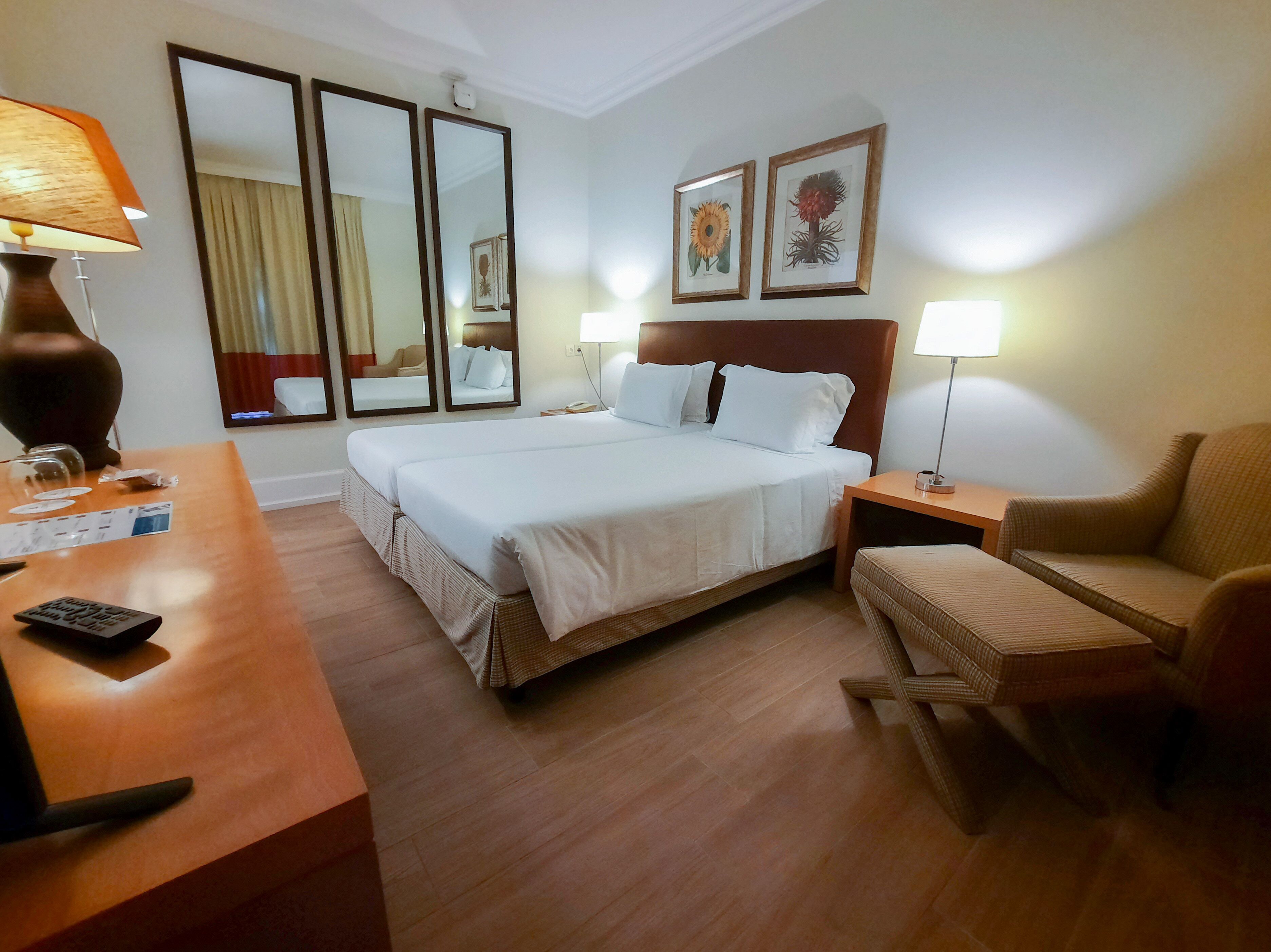 A hotel room with a large bed, two bedside tables, a chair, a desk with a lamp, and two framed pictures on the wall.