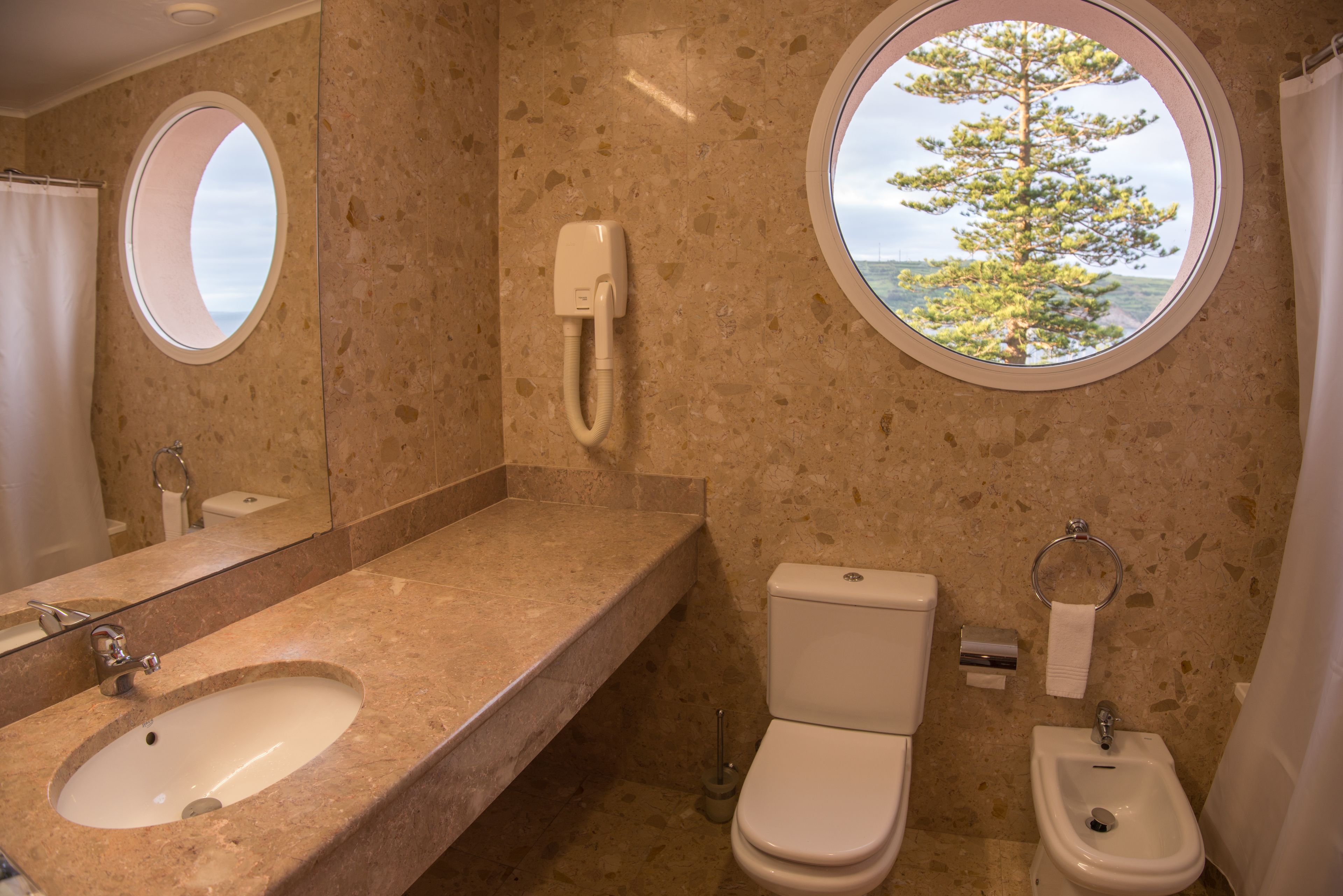 A bathroom with a marble sink, toilet, bidet, and two round windows offering views of trees and the sky.
